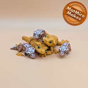 May include: A collection of five gecko figurines in various poses. The geckos are primarily yellow and brown, with blue and white patterns. The image includes a circular logo that reads "MatMire Makes Authorized Seller."
