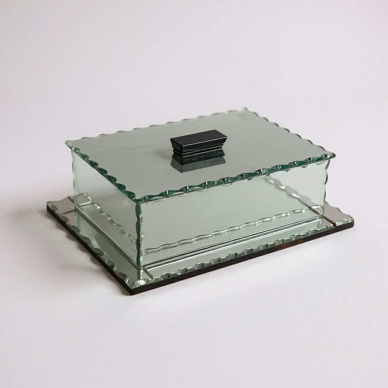 May include: A rectangular glass box with a mirrored base and lid. The lid has a small, black rectangular handle. The edges of the box and lid have a scalloped design. The box is on a white background.