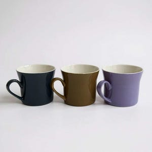 May include: Three ceramic mugs in navy, brown, and lavender, each with a cream interior. The mugs have a curved handle and a slightly flared rim. The mugs are displayed against a white background.
