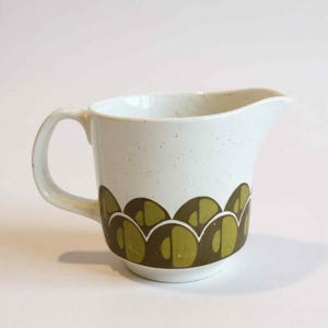 Puede incluir: Cremera de cerámica color crema con asa y boquilla. La cremera presenta un acabado moteado y un patrón repetitivo de formas verde oliva alrededor de la base. Una pieza clásica para servir crema o leche.