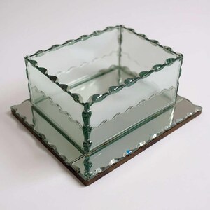 May include: A clear glass box with a scalloped edge sits on a mirrored base. The box is square and has a decorative, wavy edge. The mirrored base is rectangular with a dark brown border. The design is elegant and vintage, suitable for display.