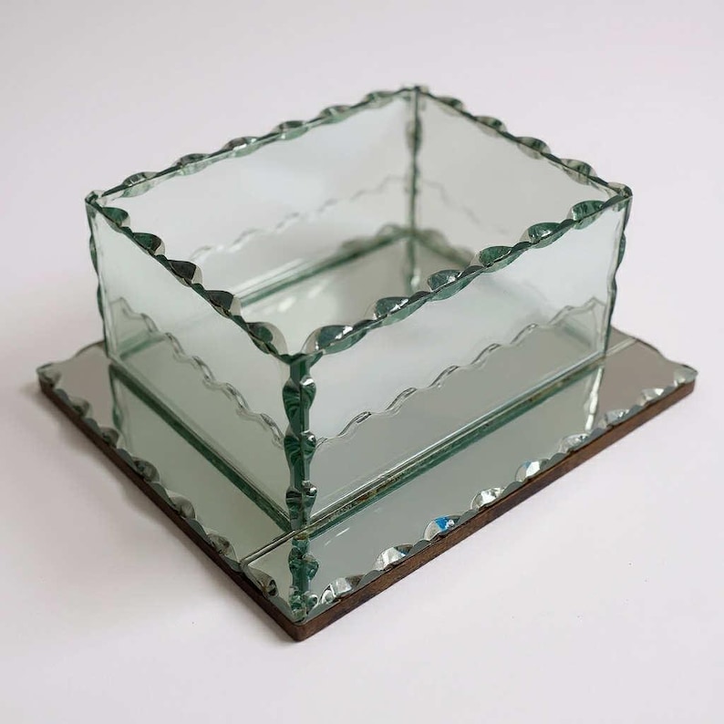 May include: A clear glass box with a scalloped edge sits on a mirrored base. The box is square and has a decorative, wavy edge. The mirrored base is rectangular with a dark brown border. The design is elegant and vintage, suitable for display.