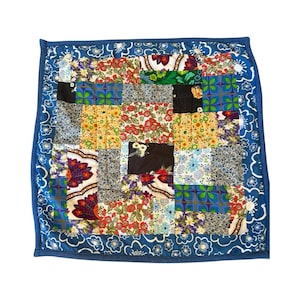 May include: Square patchwork quilt with a blue border. The quilt features a variety of floral and geometric patterns in red, yellow, blue, and black. The border is a solid blue with a white floral pattern.