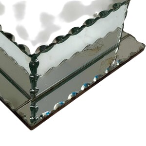 May include: A mirrored glass box with a scalloped edge. The box has a rectangular shape and is made of reflective glass. The edges are trimmed with a decorative, wavy design. The box sits on a dark brown base.