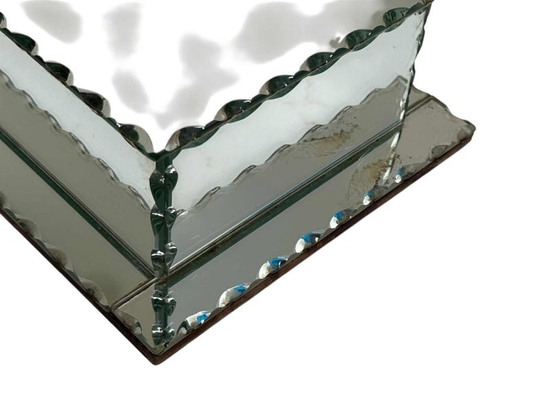 May include: A mirrored glass box with a scalloped edge. The box has a rectangular shape and is made of reflective glass. The edges are trimmed with a decorative, wavy design. The box sits on a dark brown base.