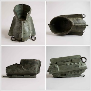 May include: Four views of a vintage, weathered metal mold, likely for casting. The mold has a dark, greenish-bronze patina, with a complex, geometric shape. It features hinged sections, handles, and a cylindrical opening. The design suggests it was used for creating a specific form.
