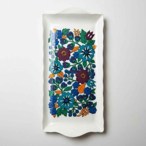 May include: A white rectangular ceramic serving tray with a floral pattern. The flowers are in shades of blue, purple, orange, and green. The tray has a scalloped edge and a small handle at the top.