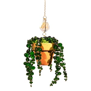 May include: A hanging planter with a small, gold-colored pot filled with green moss and cascading green beaded strands. A gold chain suspends the pot from a decorative crystal. The planter is set against a white background.