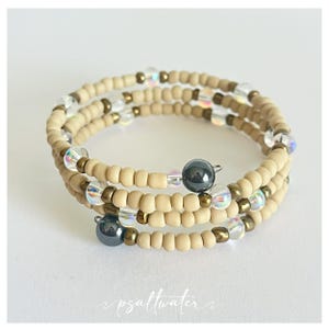 May include: A wrap bracelet featuring tan seed beads, iridescent crystals, and dark gray beads. The bracelet is multi-strand and wraps around the wrist. The design includes gold-colored metal accents. The text "psaltwater" is at the bottom.