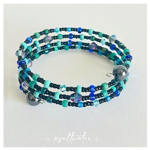 May include: A wrap bracelet with multiple strands of teal, turquoise, and blue beads, interspersed with iridescent crystals and black beads. The bracelet, with a magnetic clasp, is designed to wrap around the wrist several times.