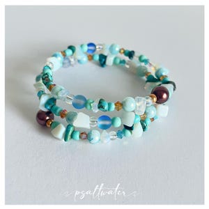 May include: A multi-strand bracelet with a spiral design. The bracelet features a variety of beads in shades of blue, turquoise, white, and clear, with accents of gold and dark brown. The beads are of various shapes and sizes, creating a textured look.