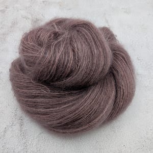 May include: A round, loosely wound skein of dark mauve yarn with a soft, fuzzy, and slightly translucent texture. The fibers are fine and delicate, creating a halo effect across the surface of the yarn ball, which is presented in a circular shape.