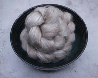 Puede incluir: Primer plano de un cuenco verde oscuro lleno de fibra trenzada gris claro. La fibra parece suave y esponjosa, con sutiles variaciones de color. El cuenco está sobre una superficie gris claro.