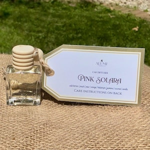 Puede incluir: Un frasco transparente y cuadrado de ambientador para coche con una tapa de madera clara y una etiqueta que dice "PINK SOLARA". La etiqueta enumera las notas de la fragancia y las instrucciones de cuidado.