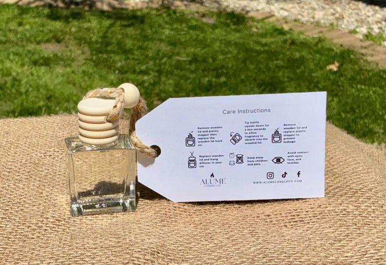 Peut inclure: Un flacon diffuseur carr&eacute; en verre transparent avec un couvercle en bois et une corde beige. Une &eacute;tiquette blanche avec des instructions d'entretien est attach&eacute;e. Le flacon est pos&eacute; sur une surface en toile de jute avec un fond herbeux vert.