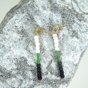 May include: A pair of drop earrings featuring gold-tone posts and dangling segments of beads. Each earring consists of four white cylindrical beads, a single green seed bead, and four black cylindrical beads arranged in a vertical line.