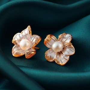 May include: A pair of flower-shaped earrings. Each earring features a central pearl surrounded by white petals with gold-colored edges and accents. The earrings are set against a dark teal background.