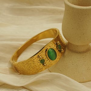 May include: A gold-colored bracelet with a textured surface and three green oval-shaped stones. The bracelet is next to a small, beige ceramic cup with a handle. The background is a cream-colored fabric.