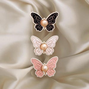 May include: Three butterfly brooches in black, white, and pink, each with a pearl center. The brooches have detailed wing designs and are set against a soft, cream-colored fabric. These decorative pins are suitable for clothing or accessories.