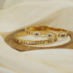May include: Two gold-colored bangles with intricate designs rest on a wooden plate. One bangle features a series of rectangular embellishments, while the other showcases an eye-shaped motif with blue and clear accents. The background is a soft, cream-colored fabric.