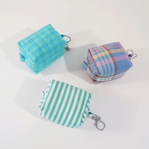 Könnte beinhalten: Drei kleine, faltbare Stofftaschen mit Schlüsselanhängern. Die Taschen haben verschiedene Muster: blau-grünes Gingham, blau-rosa Karos und grün-weiße Streifen.