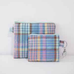 Op de afbeelding: Twee gevoerde ritszakjes met een geruit patroon in tinten blauw, roze, geel en bruin. Het kleinere zakje heeft een sleutelhanger.