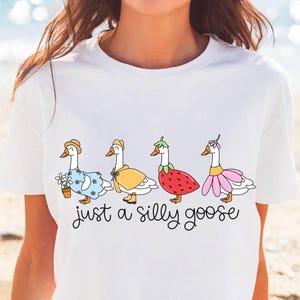 May include: White t-shirt featuring a whimsical illustration of four geese dressed in various outfits, including a dress, a cape, and a strawberry costume. The phrase "just a silly goose" is written below the geese in a playful font.