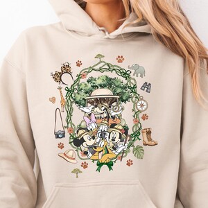 Peut inclure: Sweat &agrave; capuche beige avec un motif color&eacute; de Mickey, Donald, Dingo et Daisy en tenue de safari. Le design comprend des &eacute;l&eacute;ments de safari comme une boussole, des jumelles et une girafe, entour&eacute;s d'une guirlande de vigne verte.