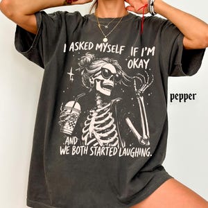 May include: Dark gray t-shirt featuring a skeleton graphic wearing sunglasses and holding a drink. The text on the shirt reads "I asked myself if I'm okay, and we both started laughing."