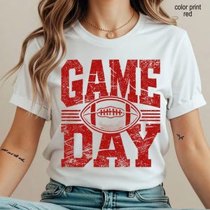 May include: A white short-sleeved t-shirt featuring a distressed red graphic print. The design displays the text "GAME DAY" in large block letters with a football illustration centered between the words. The text "color print red" is visible.