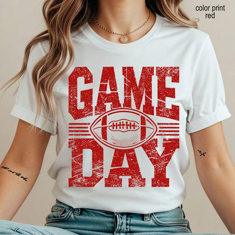 May include: A white short-sleeved t-shirt featuring a distressed red graphic print. The design displays the text "GAME DAY" in large block letters with a football illustration centered between the words. The text "color print red" is visible.