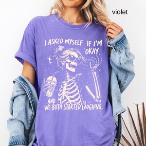 May include: Violet-colored t-shirt with a skeleton graphic wearing sunglasses and holding a drink. The text on the shirt reads, "I asked myself if I'm okay, and we both started laughing."