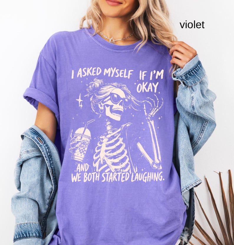 May include: Violet-colored t-shirt with a skeleton graphic wearing sunglasses and holding a drink. The text on the shirt reads, "I asked myself if I'm okay, and we both started laughing."