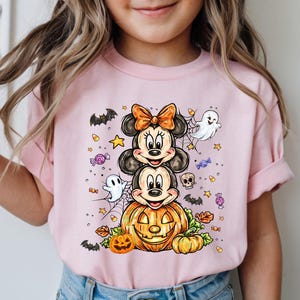 Puede incluir: Camiseta rosa de manga corta con un dise&ntilde;o de Halloween. El gr&aacute;fico presenta personajes de dibujos animados apilados, una calabaza tallada, fantasmas, murci&eacute;lagos, estrellas y caramelos. Los personajes tienen orejas negras y lazos naranjas.