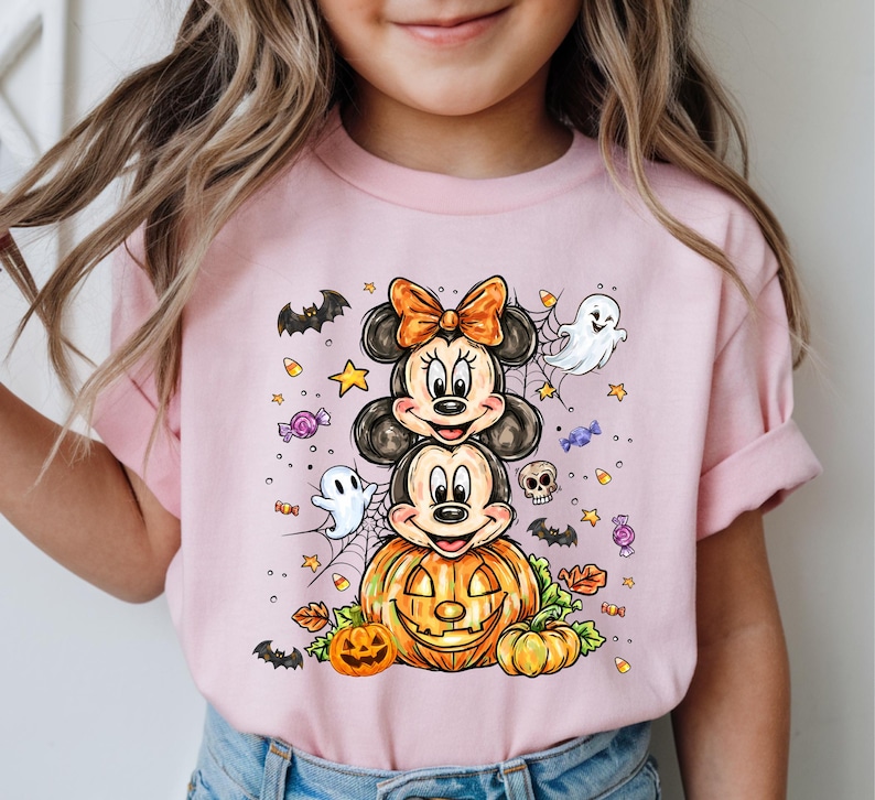 Puede incluir: Camiseta rosa de manga corta con un dise&ntilde;o de Halloween. El gr&aacute;fico presenta personajes de dibujos animados apilados, una calabaza tallada, fantasmas, murci&eacute;lagos, estrellas y caramelos. Los personajes tienen orejas negras y lazos naranjas.