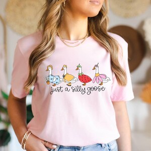 May include: A light pink t-shirt featuring a whimsical illustration of four geese dressed in various outfits. The geese are adorned with a flower pot, a yellow dress, a strawberry, and a pink flower. The text "just a silly goose" is below the geese.