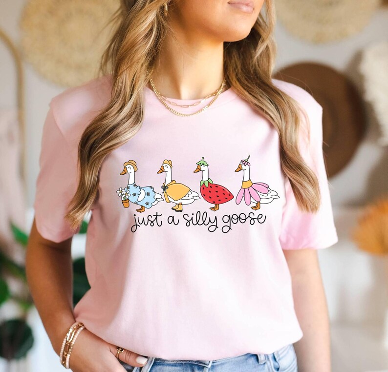 May include: A light pink t-shirt featuring a whimsical illustration of four geese dressed in various outfits. The geese are adorned with a flower pot, a yellow dress, a strawberry, and a pink flower. The text "just a silly goose" is below the geese.