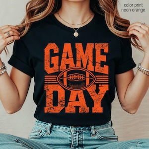 May include: A black short-sleeved t-shirt featuring the text "GAME DAY" in a distressed neon orange font. Centered between the words is a graphic of an American football. The design is printed in a bold, textured style on the front of the shirt.