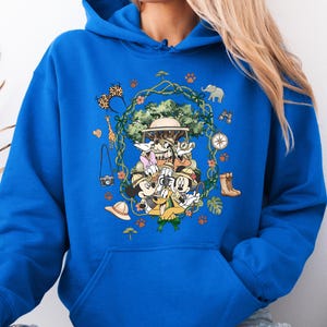 Peut inclure: Sweat &agrave; capuche bleu roi avec un motif sur le th&egrave;me du safari. Le design comprend Mickey Mouse, Donald Duck, Dingo et d'autres personnages en tenue de safari, entour&eacute;s d'ic&ocirc;nes li&eacute;es au safari comme une boussole, des jumelles et un appareil photo. Le motif est centr&eacute; sur le devant.
