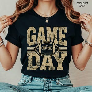 May include: A black short-sleeved t-shirt featuring the text GAME DAY in a distressed, sand-colored font. Centered between the words is a graphic illustration of an American football. The design has a weathered, vintage-inspired texture.