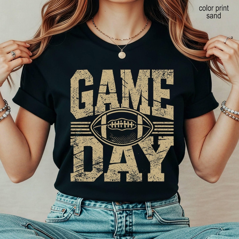May include: A black short-sleeved t-shirt featuring the text GAME DAY in a distressed, sand-colored font. Centered between the words is a graphic illustration of an American football. The design has a weathered, vintage-inspired texture.