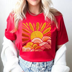 May include: A red t-shirt featuring an embroidered design of a yellow sun with orange rays and clouds. The sun design is centered on the shirt. The shirt is made of a soft material.