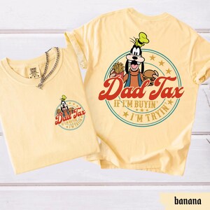 May include: Two banana-colored t-shirts. One shirt has a small graphic on the front with the text "Dad Tax" and a cartoon character. The other shirt has a larger graphic on the back with the same text and character.