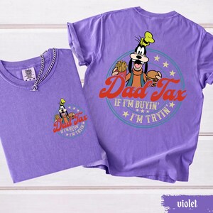 May include: Two violet-colored t-shirts. One shirt has a small graphic of Goofy with the words "Dad Tax" and "If I'm buyin' I'm tryin'". The other shirt has the same graphic on the back. A silver chain necklace is on the front of one shirt.