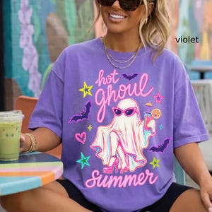 May include: A violet t-shirt with a graphic of a ghost wearing sunglasses, high heels, and holding a cocktail. The text "hot ghoul summer" is written in pink script. Bats, stars, and hearts surround the ghost.