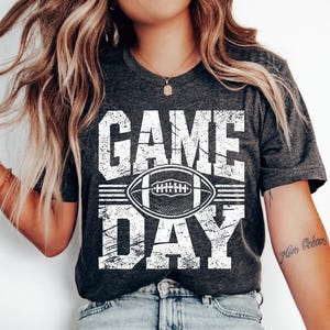 Puede incluir: Camiseta gris oscuro con las palabras "GAME DAY" en letras blancas desgastadas, con un gr&aacute;fico de f&uacute;tbol americano blanco en el centro. La camiseta es de estilo casual, adecuada para los fan&aacute;ticos del deporte.
