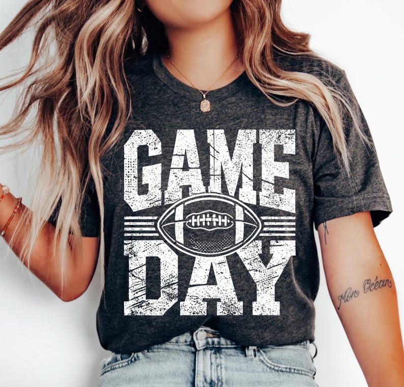 Puede incluir: Camiseta gris oscuro con las palabras "GAME DAY" en letras blancas desgastadas, con un gr&aacute;fico de f&uacute;tbol americano blanco en el centro. La camiseta es de estilo casual, adecuada para los fan&aacute;ticos del deporte.