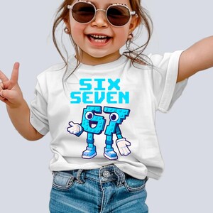 May include: White t-shirt with the words "SIX SEVEN" in pixelated blue letters. Below the text are the numbers 6 and 7 designed as cartoon characters in blue with white gloves and shoes. The shirt is worn by a child.