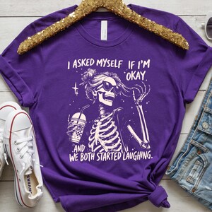 May include: A purple t-shirt featuring a skeleton graphic wearing sunglasses and holding a beverage. The text on the shirt reads, "I asked myself if I'm okay, and we both started laughing."