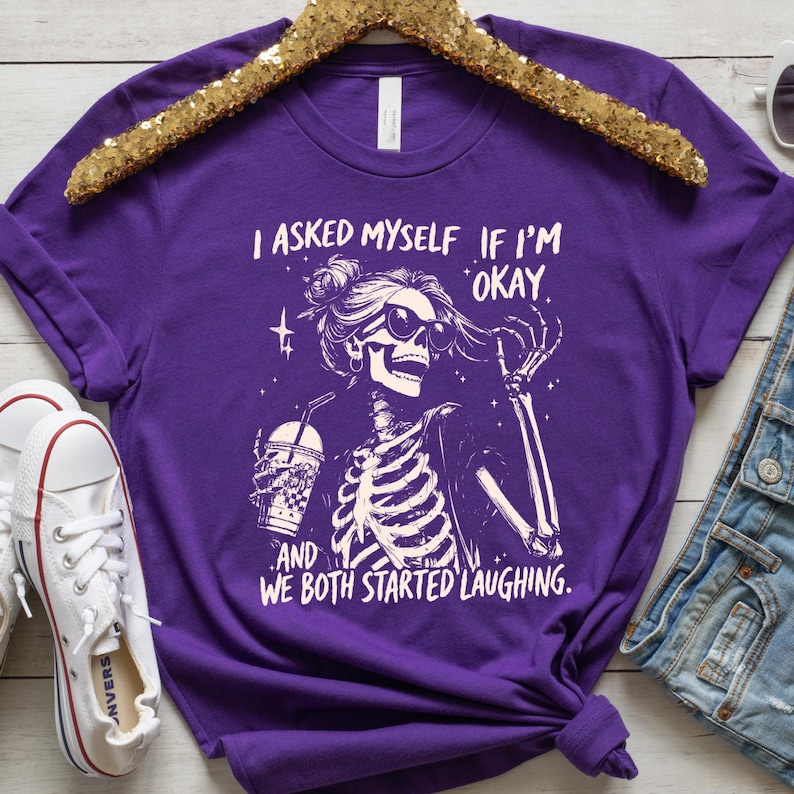 May include: A purple t-shirt featuring a skeleton graphic wearing sunglasses and holding a beverage. The text on the shirt reads, "I asked myself if I'm okay, and we both started laughing."
